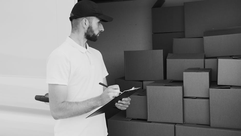 A man wearing a white shirt and jeans operates outside a residential property during a home relocation, preparing to load cardboard boxes into the open rear door of a white moving van parked on the st