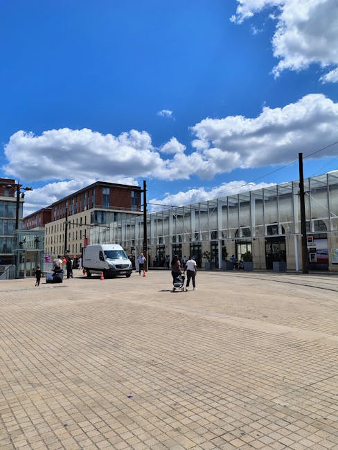 A wide shot of an outdoor area with a large, paved open space under a bright blue sky filled with scattered white clouds. In the background, there are modern multi-storey buildings with glass and brick facades, and a long, transparent structure resembling a glass corridor or entrance. Several people are present; some are carrying or moving items, while others are standing or walking. A white panel van is parked near the building, with a few orange traffic cones nearby. The scene suggests a home relocation or furniture transport process, with the activity organized around loading or unloading items, consistent with professional removals services such as those offered by Twickenham Man and Van.