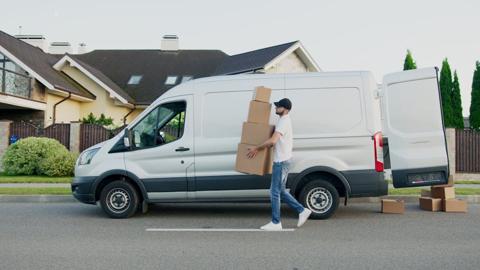 A man wearing a black cap, white t-shirt, and blue jeans is standing on the street next to a white cargo van, which is parked in front of residential houses with cream-colored walls, dark rooftops, and large windows. The man is carrying a stack of three cardboard moving boxes, with one more box on the ground beside him. Additional cardboard boxes are visible on the pavement near the open rear door of the van, which is in the loading process during a home relocation. The van's sliding side door is closed, and the rear doors are open, revealing the empty cargo space. The scene is set during daylight hours with bright, natural lighting, and the front of the van faces the left side of the image. The surroundings include a well-maintained lawn, a wooden fence, and tall green trees, indicating a suburban neighbourhood where [COMPANY_NAME] likely provides removals and furniture transport services related to house moves near Twickenham Stadium, Twickenham, as described on the web page.